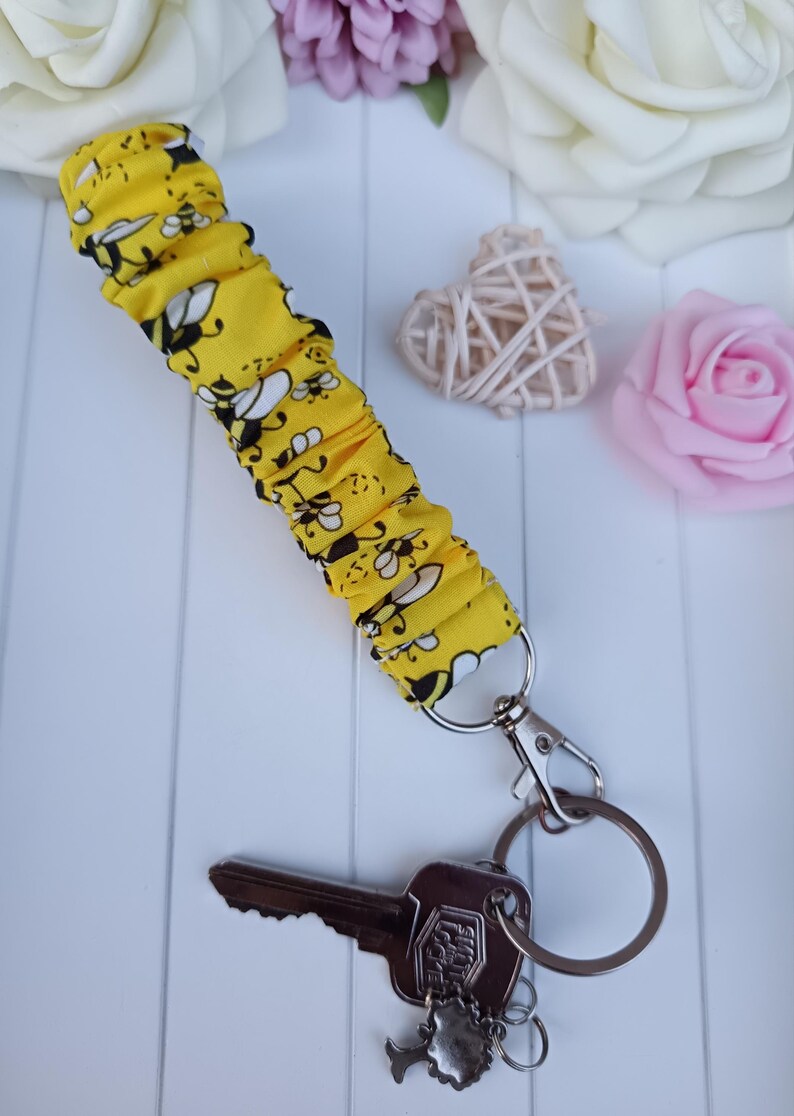 Pode incluir: Porta-chaves de tecido amarelo com um padr&atilde;o de abelhas, preso a um porta-chaves e fecho prateados. Uma chave com um pendente decorativo tamb&eacute;m est&aacute; presa. O fundo inclui flores decorativas e um objeto em forma de cora&ccedil;&atilde;o tecido.