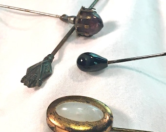 May include: Four vintage hat pins with different colored glass and metal accents. The pins are on a white fabric background.