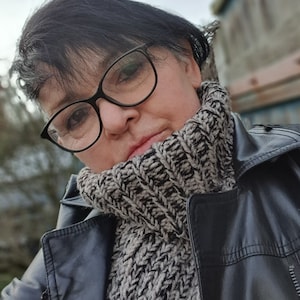 Könnte beinhalten: Eine Frau trägt eine schwarze Lederjacke mit einem dicken, grau-schwarzen Strickrollkragen. Sie trägt eine schwarze Brille.