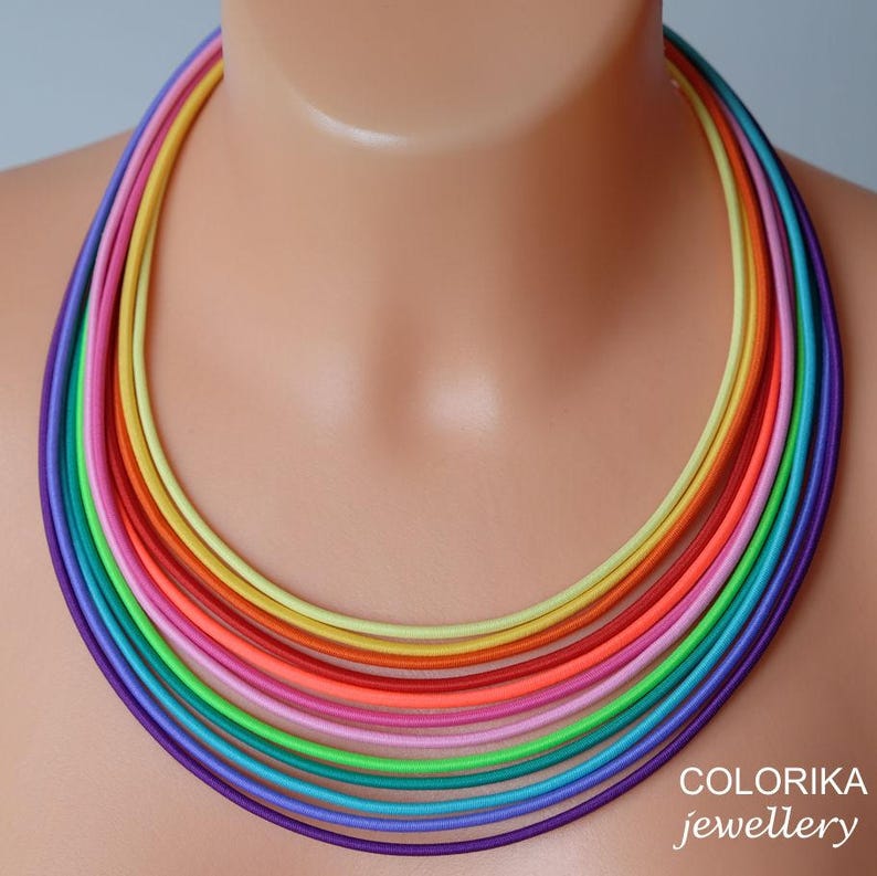 May include: A multi-coloured rainbow necklace made of thin, layered cords. The necklace is made of purple, blue, teal, green, lime green, pink, orange, red, yellow, and light yellow cords.