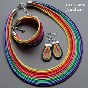 May include: A rainbow-coloured necklace, bracelet and earrings set. The necklace is made of multiple strands of colourful cord, and the bracelet is a single strand of cord. The earrings are teardrop-shaped and have a silver hook.