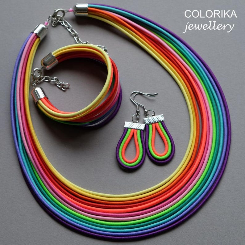 May include: A rainbow-coloured necklace, bracelet and earrings set. The necklace is made of multiple strands of colourful cord, and the bracelet is a single strand of cord. The earrings are teardrop-shaped and have a silver hook.