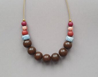 Puede incluir: Un collar de cuentas de madera marrón con cuentas rojas, rosadas y azules. El collar está encordado en un cordón dorado.