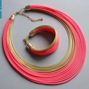 Pu&ograve; includere: Un vivace set di gioielli con una collana a pi&ugrave; fili e un braccialetto abbinato. La collana e il braccialetto combinano cordoncini rosa acceso con dettagli in metallo dorato. Il set &egrave; esposto su uno sfondo grigio, mettendo in risalto la sua audace combinazione di colori.