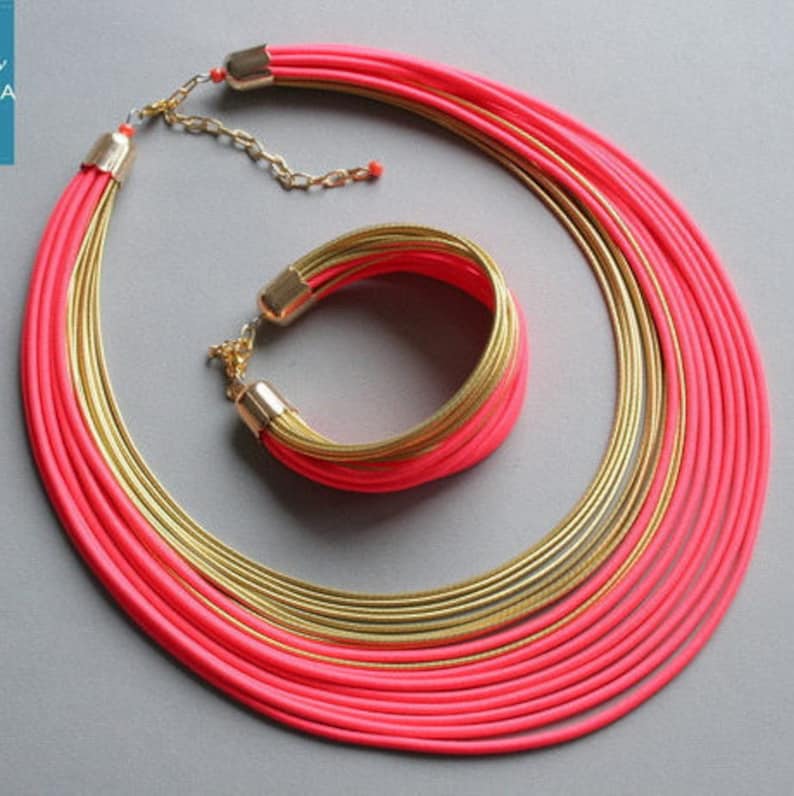 Pu&ograve; includere: Un vivace set di gioielli con una collana a pi&ugrave; fili e un braccialetto abbinato. La collana e il braccialetto combinano cordoncini rosa acceso con dettagli in metallo dorato. Il set &egrave; esposto su uno sfondo grigio, mettendo in risalto la sua audace combinazione di colori.