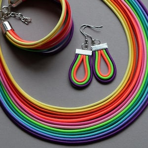 May include: A rainbow-coloured necklace and bracelet set with matching earrings. The necklace and bracelet are made of multiple strands of elastic cord in various colours, including red, orange, yellow, green, blue, indigo, and violet. The earrings are teardrop-shaped and feature a similar rainbow design.