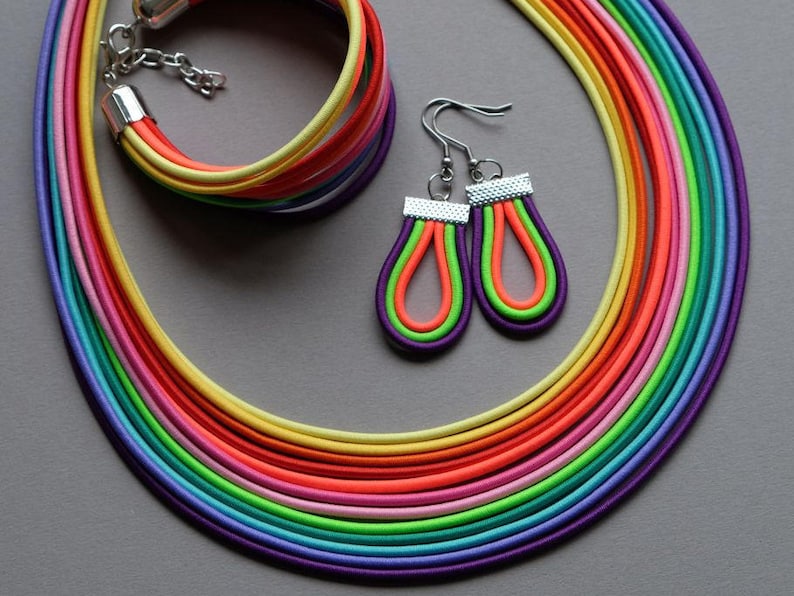 May include: A rainbow-coloured necklace and bracelet set with matching earrings. The necklace and bracelet are made of multiple strands of elastic cord in various colours, including red, orange, yellow, green, blue, indigo, and violet. The earrings are teardrop-shaped and feature a similar rainbow design.