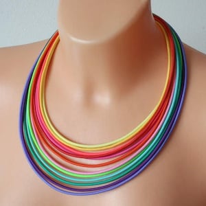 Puede incluir: Un collar multicolor de múltiples hebras. El collar está compuesto por numerosos cordones finos y flexibles en tonos morado, azul, verde, amarillo, naranja y rosa. El collar se muestra en un maniquí.
