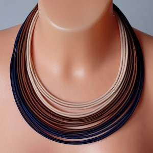 Puede incluir: Un collar de múltiples capas con un esquema de color degradado. El collar está hecho de tela y presenta tonos de azul marino, marrón y beige.