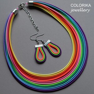 May include: A rainbow coloured necklace and matching earrings. The necklace is made of multiple strands of elastic cord in various colours, including red, orange, yellow, green, blue, indigo, and violet. The earrings are teardrop shaped and feature the same rainbow colours.