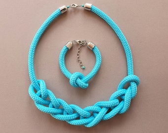 Puede incluir: Un conjunto de collar y pulsera de cuerda azul turquesa con cierres plateados. El collar tiene un diseño de nudo grande y la pulsera tiene un diseño de nudo más pequeño.
