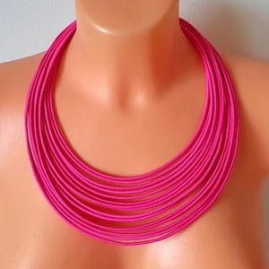 Könnte beinhalten: Eine mehrsträngige Halskette mit zahlreichen dünnen, leuchtend pinkfarbenen Kordeln, die in einem kreisförmigen, geschichteten Design eng am Hals anliegen. Die Kordeln sind in Farbe und Textur einheitlich und erzeugen eine markante, gestapelte Optik.
