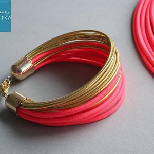Pu&ograve; includere: Un vivace set di bracciale e collana. Il bracciale presenta pi&ugrave; fili di metallo color oro e tessuto rosa brillante, fissati con terminali dorati e una catena. La collana ha un design simile. Il marchio "handmade by COLORIKA" &egrave; nell'angolo in alto a sinistra.