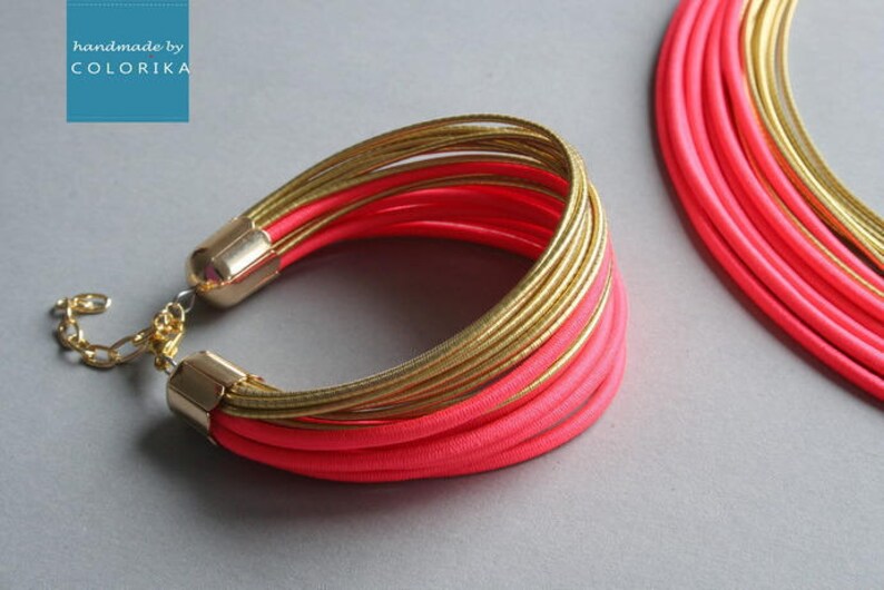 Pu&ograve; includere: Un vivace set di bracciale e collana. Il bracciale presenta pi&ugrave; fili di metallo color oro e tessuto rosa brillante, fissati con terminali dorati e una catena. La collana ha un design simile. Il marchio "handmade by COLORIKA" &egrave; nell'angolo in alto a sinistra.