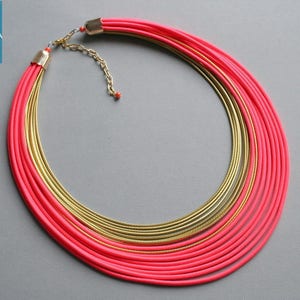 Pu&ograve; includere: Collana a pi&ugrave; strati con elementi rosa acceso e dorati. La collana presenta pi&ugrave; fili di materiale rosa e anelli in metallo color oro. Il testo "handmade by COLORIKA" &egrave; in un quadrato blu nell'angolo in alto a sinistra.