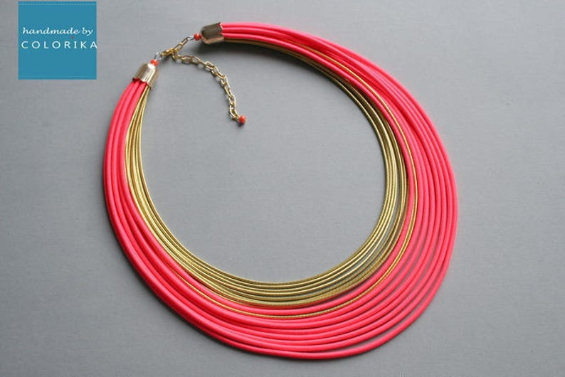Pu&ograve; includere: Collana a pi&ugrave; strati con elementi rosa acceso e dorati. La collana presenta pi&ugrave; fili di materiale rosa e anelli in metallo color oro. Il testo "handmade by COLORIKA" &egrave; in un quadrato blu nell'angolo in alto a sinistra.