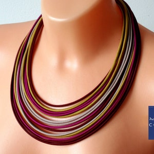 May include: A multi-strand necklace in shades of burgundy, magenta, olive green, and grey. The necklace is made up of multiple strands of smooth, rounded material, creating a layered effect. The necklace is displayed on a mannequin.