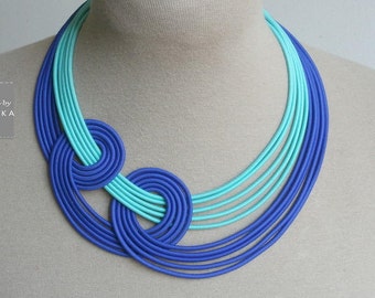 May include: A multi-layered necklace made of blue and turquoise fabric. The necklace is arranged in a circular pattern with a knot in the center.