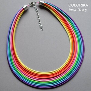 May include: A multi-coloured rainbow necklace made of thin, layered cords. The necklace is made of purple, blue, green, turquoise, lime green, pink, orange, yellow, and light yellow cords. The necklace has a silver clasp.