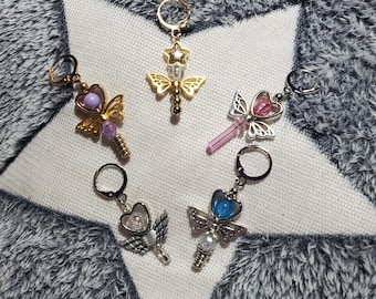 May include: Five decorative keychains with celestial and butterfly designs. The keychains feature gold, silver, and pearl accents, with colorful gems and star details. The keychains are arranged on a white star-shaped surface.