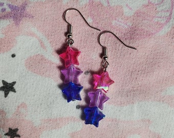May include: A pair of dangle earrings with three star-shaped beads. The stars are pink, purple, and blue.