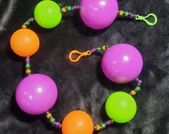 May include: A toy consisting of two purple, two lime green, and two orange plastic spheres strung together with small, multi-colored beads. The string features two bright orange plastic clips at the ends for attachment, creating a circular loop.
