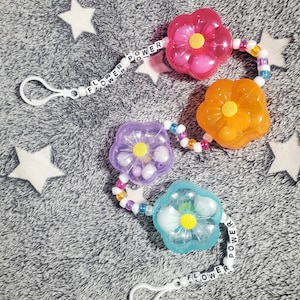 Puede incluir: Un conjunto de cuatro clips para chupete con forma de flor en rosa, naranja, morado y azul. Cada flor tiene un centro amarillo y está conectada por una cadena de cuentas. Las palabras "FLOWER POWER" están escritas en cuentas blancas.