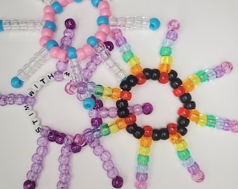 Może przedstawiać: Trzy kolorowe bransoletki przyjaźni wykonane z małych plastikowych koralików. Jedna bransoletka jest fioletowa z napisem "STIM", druga jest tęczowa, a trzecia jest różowa i niebieska.
