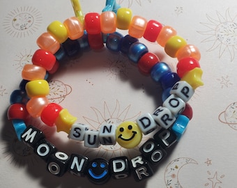 Może przedstawiać: Kolorowa bransoletka z koralików z napisami "Moon Drop" i "Sun Drop" wyłożonymi czarnymi koralikami. Bransoletka ma dwa koraliki z uśmiechami.