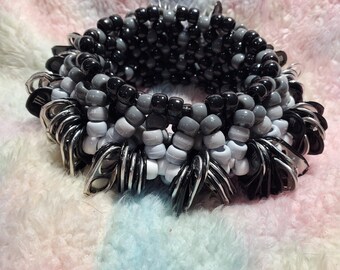 May include: A handmade bracelet featuring a layered design of black, gray, and white beads. The bracelet is accented with silver-colored metal rings, creating a unique and textured accessory. The bracelet is shown on a soft, multi-colored surface.