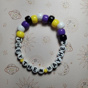 Könnte beinhalten: Ein Armband aus Perlen mit gelben, schwarzen, weißen und violetten Perlen. Die Perlen buchstabieren "They Them May He" in schwarzen Buchstaben.