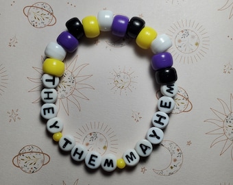 May include: A beaded bracelet with yellow, black, white, and purple beads. The beads spell out "They Them May He" in black lettering.