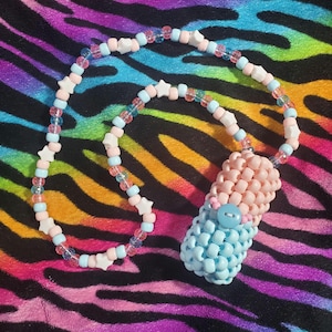 May include: A beaded necklace featuring a cylindrical pendant made of woven light pink and light blue plastic pony beads. The necklace strand consists of alternating light pink, light blue, and clear beads interspersed with white star-shaped beads.