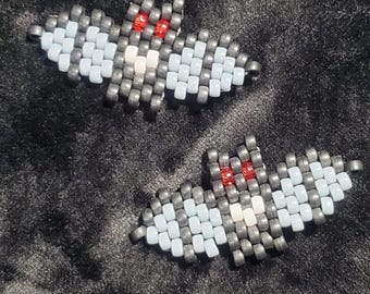 May include: Two bat-shaped beaded ornaments crafted from small grey, light blue, and white beads. Each bat features two prominent red beads for eyes, with a central white bead on the chest and grey beads forming the wings and body structure.