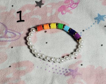 May include: A rainbow colored beaded bracelet with white pearl beads. The bracelet is on a pink and white patterned fabric.