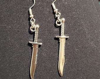 May include: A pair of silver earrings shaped like a knife. The earrings are hanging from a black surface.