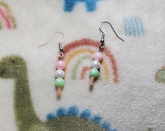 May include: A pair of beaded earrings with a pastel color scheme. Each earring features a silver hook and a vertical arrangement of pink, white, and green beads, with a cluster of smaller pink beads at the bottom. The earrings are displayed on a white and pastel-colored dinosaur-themed fabric.