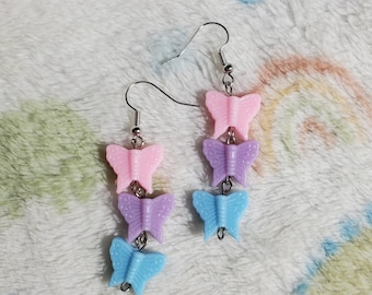 May include: A pair of dangle earrings featuring three butterfly charms in shades of pink, purple, and blue.
