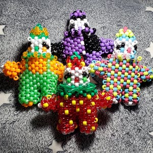 Könnte beinhalten: Vier Figuren aus Perlen in verschiedenen Farben, darunter Orange, Grün, Rot, Lila und Regenbogenfarben. Jede Figur hat ein eigenes Design, einige mit Hüten und Kragen. Die Figuren sind auf einer grauen Oberfläche mit Sternenmuster angeordnet.