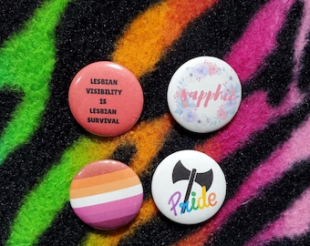 May include: Four round buttons with pride-themed designs. One button reads "Lesbian Visibility is Lesbian Survival" in red. Another features the word "Sapphie" in pink script surrounded by flowers. The third has the lesbian flag colors. The last has an axe and the word "Pride".