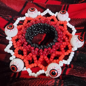 Op de afbeelding: Een cirkelvormige kralenarmband met een centrale ring van zwarte kralen omringd door concentrische lagen van rode en oranje kralen. De buitenrand is geaccentueerd met vijf witte bolvormige kralen met rode irissen en zwarte pupillen.