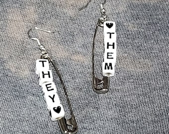 May include: A pair of silver safety pin earrings with white beads spelling out "THEY" and "THEM" with a black heart bead at the end of each word.