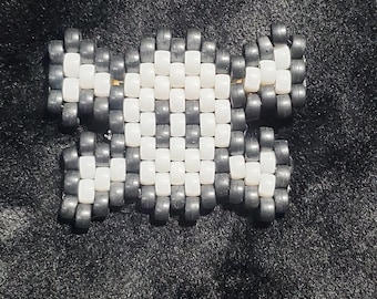May include: A skull and crossbones design crafted from small, cylindrical plastic beads. The central skull shape is composed of white beads with dark gray beads forming the outline and the crossbones extending from the sides of the skull.