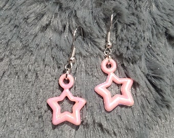 May include: A pair of pink star-shaped earrings with a silver hook. The stars have a hollow center and a shiny finish.