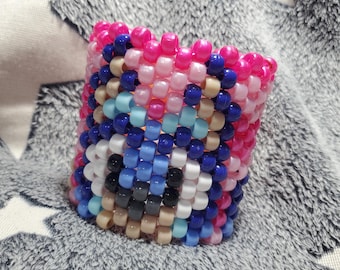 May include: A wide cuff bracelet constructed from small plastic pony beads in a grid pattern. The design features a stylized character face in shades of light blue, white, black, and tan, bordered by rows of vibrant hot pink and dark blue beads.