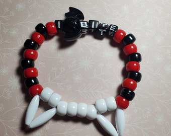 Könnte beinhalten: Ein schwarzes und rotes Perlenarmband mit einem weißen Plastik-Fledermaus-Anhänger und dem Wort "BITE" in weißen Buchstaben auf schwarzen Perlen.