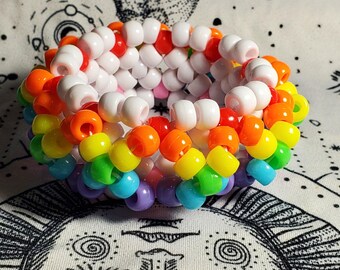 May include: A rainbow kandi bracelet made with colorful plastic beads. The bracelet is made with white, orange, yellow, green, blue, and purple beads.