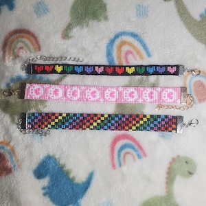 May include: Three beaded bracelets with colorful patterns. The top bracelet has a rainbow heart design, the middle bracelet has a pink and white pattern, and the bottom bracelet has a rainbow checkered pattern.