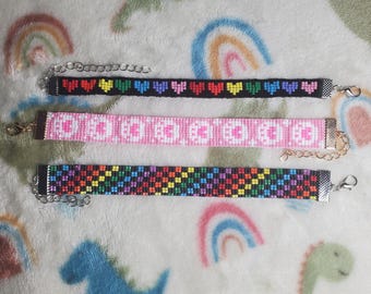 May include: Three beaded bracelets with colorful patterns. The top bracelet has a rainbow heart design, the middle bracelet has a pink and white pattern, and the bottom bracelet has a rainbow checkered pattern.