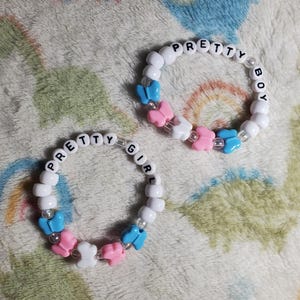 Puede incluir: Dos pulseras de cuentas con las palabras "Pretty Girl" y "Pretty Boy" escritas en ellas. Las pulseras son blancas con abalorios de mariposas rosas y azules.